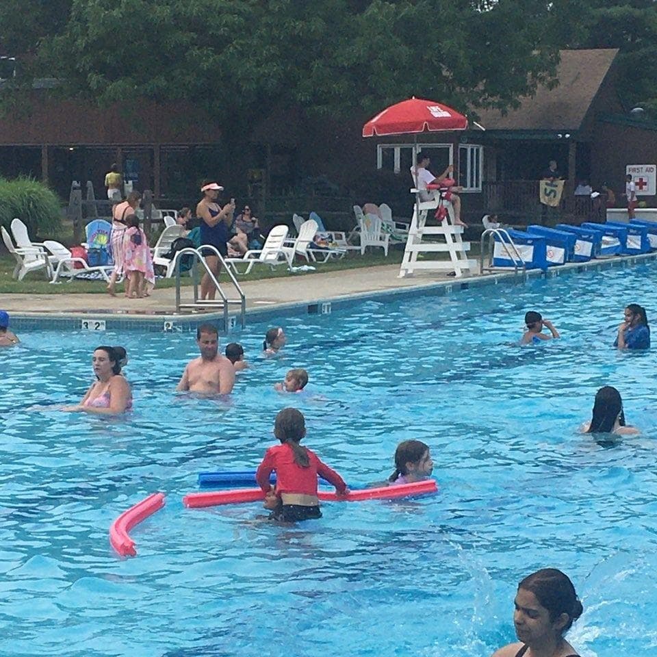 Families swimming with pool noodles while a lifeguard watches from a stand under a red umbrella.