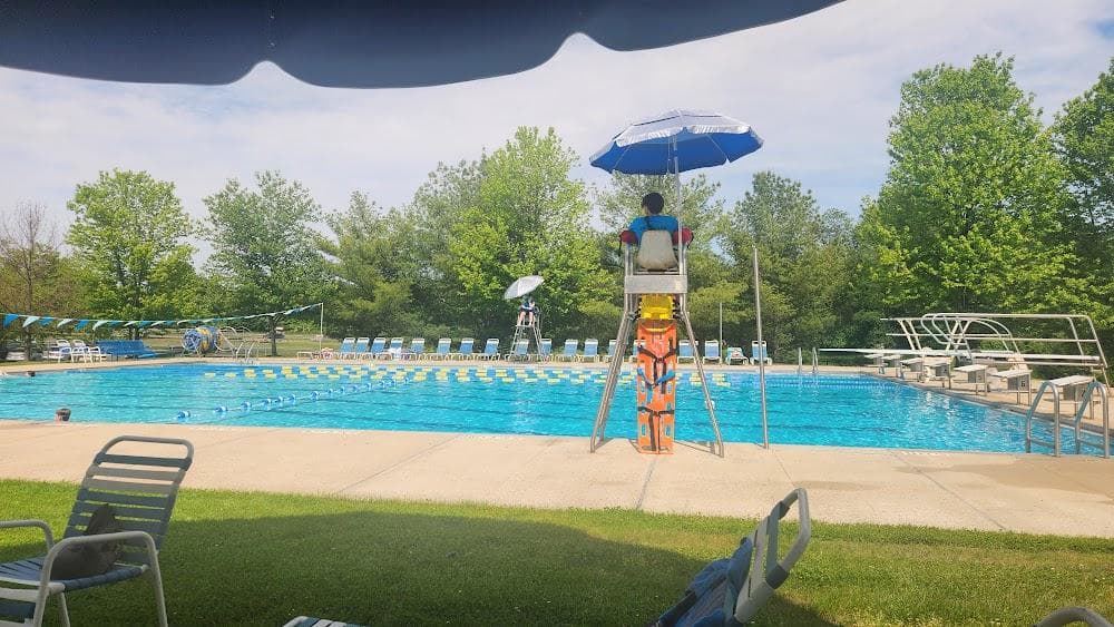 A lifeguard under a blue umbrella on a stand beside a lap pool with lane lines, seen from the lawn.